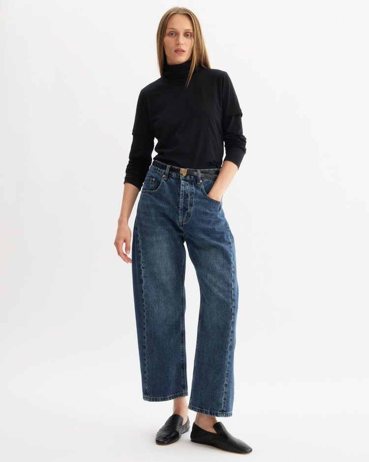 Against a white background, a person wears the Arlo Mott Prince Long Sleeve Turtleneck in Black, paired with loose blue jeans, a black belt, and black slip-on shoes. One hand is in their pocket while the other rests by their side.