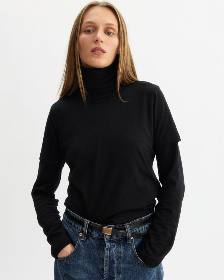 A woman with straight, light brown hair wears the Arlo Mott Prince Long Sleeve Turtleneck in Black and blue jeans. Standing against a plain white background with hands in her pockets, she looks confidently at the camera.