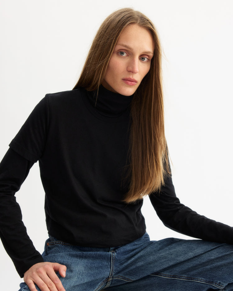 A person with long straight hair, wearing the Arlo Mott Prince Long Sleeve Turtleneck in Black under a black T-shirt and blue jeans, sits with one leg up against a plain white background.