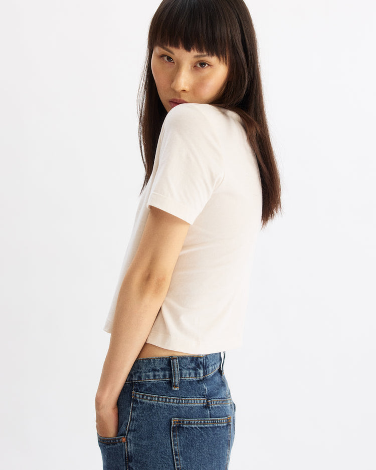 A person with straight dark hair and bangs wears the Arlo Mott Willow Short Sleeve Tee in Chalk, paired with blue jeans. They stand sideways with hands in pockets, glancing over their shoulder against a plain white background.