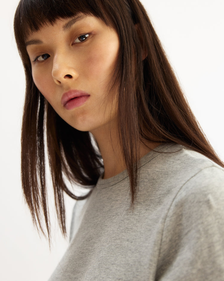 A person with straight, long brown hair and blunt bangs gazes at the camera, wearing the Arlo Mott Willow Short Sleeve Tee in Heather Grey against a plain light background.