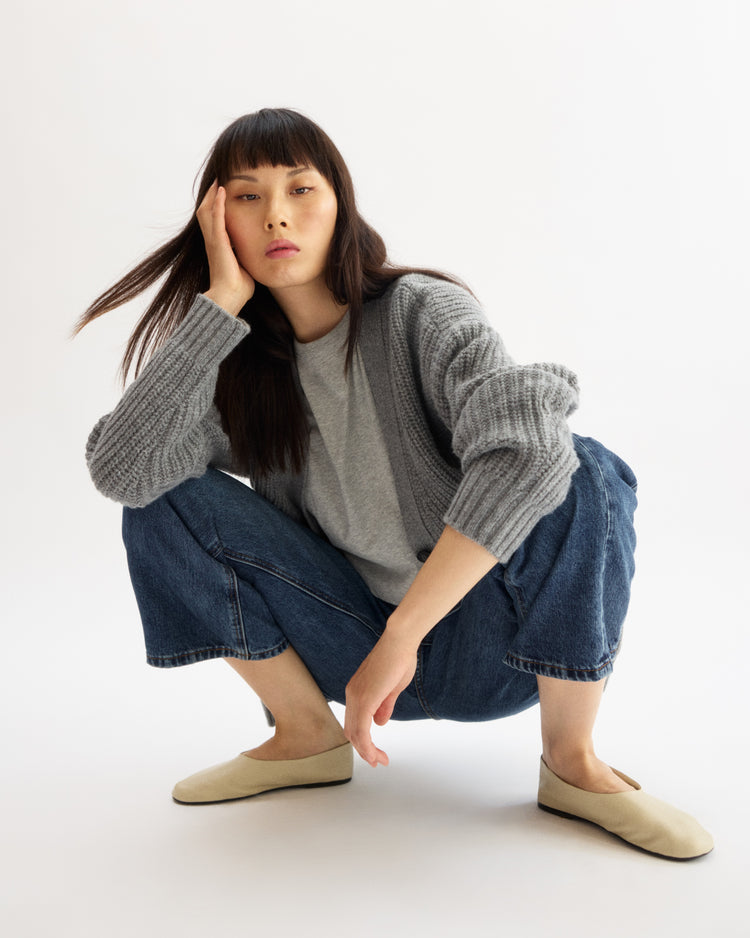 A woman with straight dark hair squats on the floor, resting her head on her hand. She wears the Arlo Mott Willow Short Sleeve Tee in Heather Grey, cropped blue jeans, and beige flats against a plain white background.