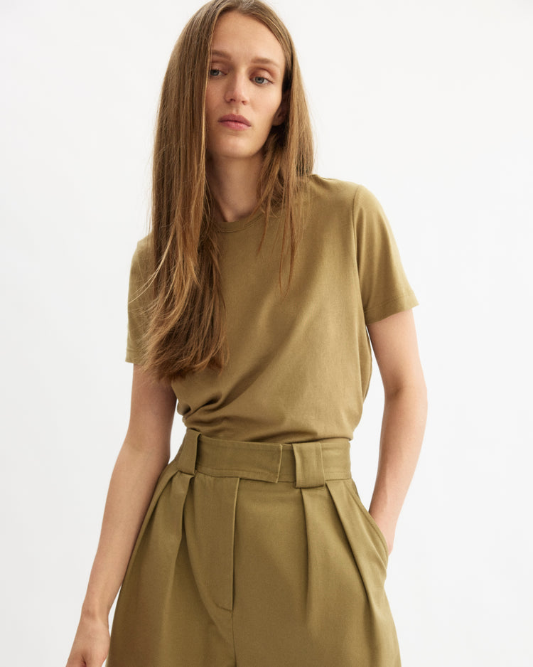 A woman with long straight hair wears the Arlo Mott Willow Short Sleeve Tee in Khaki, made in New York, tucked into high-waisted pleated trousers, standing against a plain white background.
