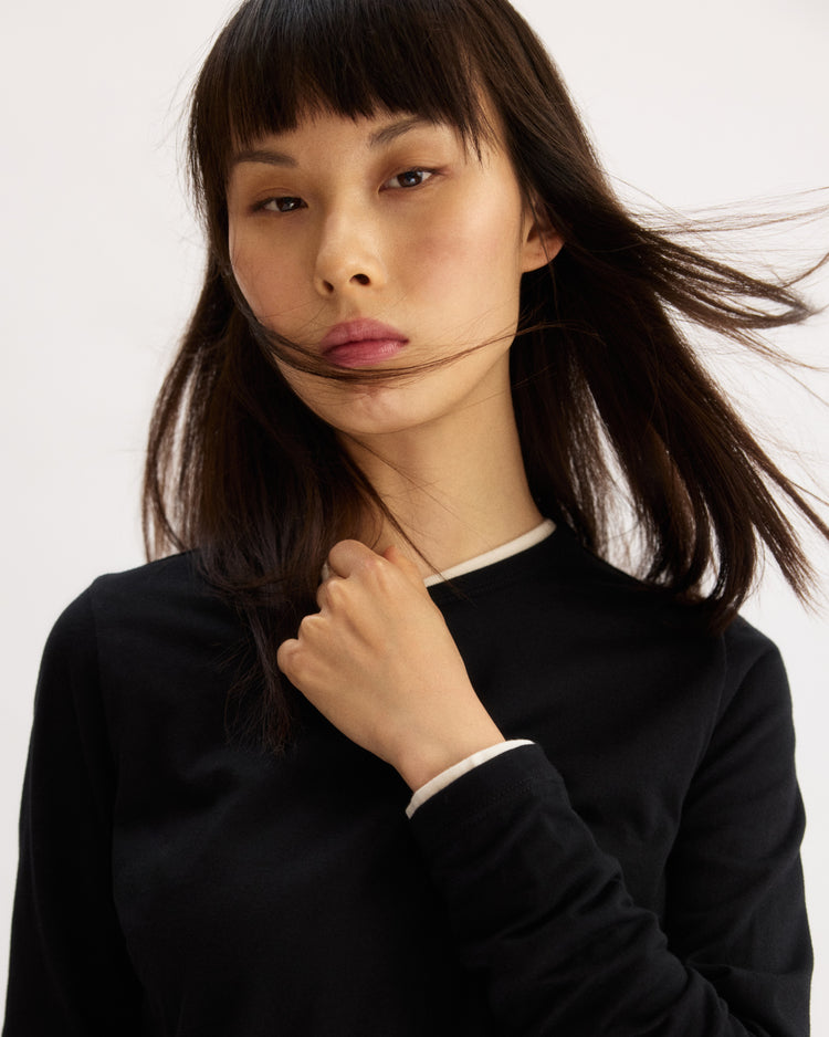 A woman with straight dark hair and bangs wears the Arlo Mott Sullivan Long Sleeve Tee in Black with white trim, looking at the camera while holding her collar. Her hair partly covers her face, and she maintains a neutral expression.
