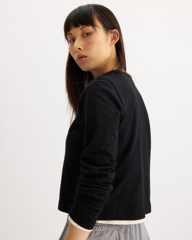 A woman with long dark hair and bangs stands against a light background, looking over her shoulder. She wears the Arlo Mott Sullivan Long Sleeve Tee in Black with a black and white striped pleated skirt, blending comfort and style.
