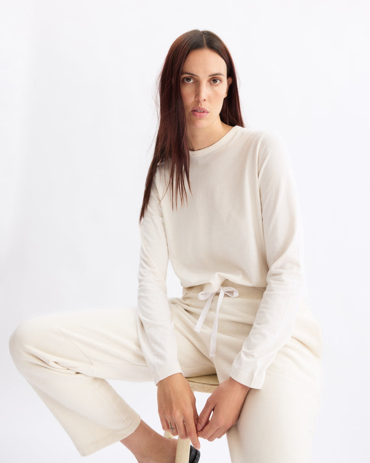 A woman with long brown hair wears the Arlo Mott Sullivan Long Sleeve Tee in Chalk and matching drawstring pants while seated on a stool against a plain white background.