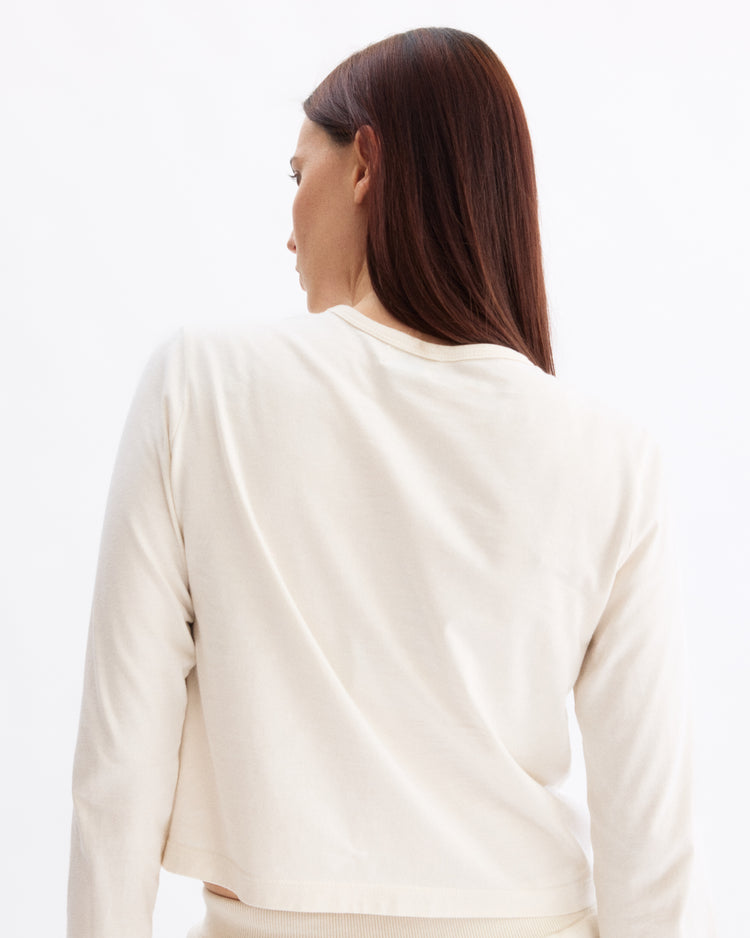 A woman with long, straight brown hair, seen from behind, models the Arlo Mott Sullivan Long Sleeve Tee in Chalk—an off-white top crafted from soft Supima cotton—against a plain white background, her face shown in profile.