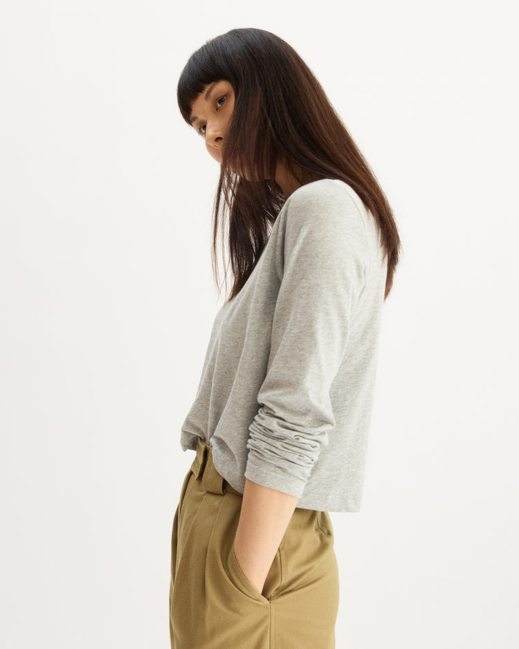 A woman with straight dark hair stands sideways, gazing ahead. She wears the Arlo Mott Sullivan Long Sleeve Tee in Heather Grey and tan high-waisted pants, hands in pockets before a plain white background.