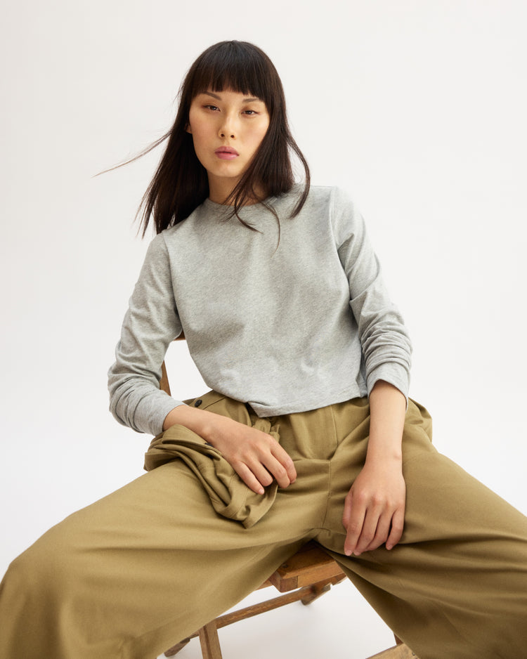 A woman with straight dark hair and bangs sits on a wooden chair against a plain background, wearing the Arlo Mott Sullivan Long Sleeve Tee in Heather Grey and wide-legged khaki pants, looking confidently at the camera. Made in Brooklyn.