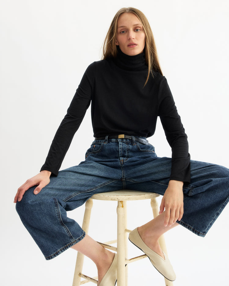 A woman with long straight hair sits on a stool against a white background, wearing the Arlo Mott Prince Long Sleeve Turtleneck in Black, wide-leg blue jeans, and beige flats. She looks relaxed with her hands on her knees—a touch of Brooklyn style.