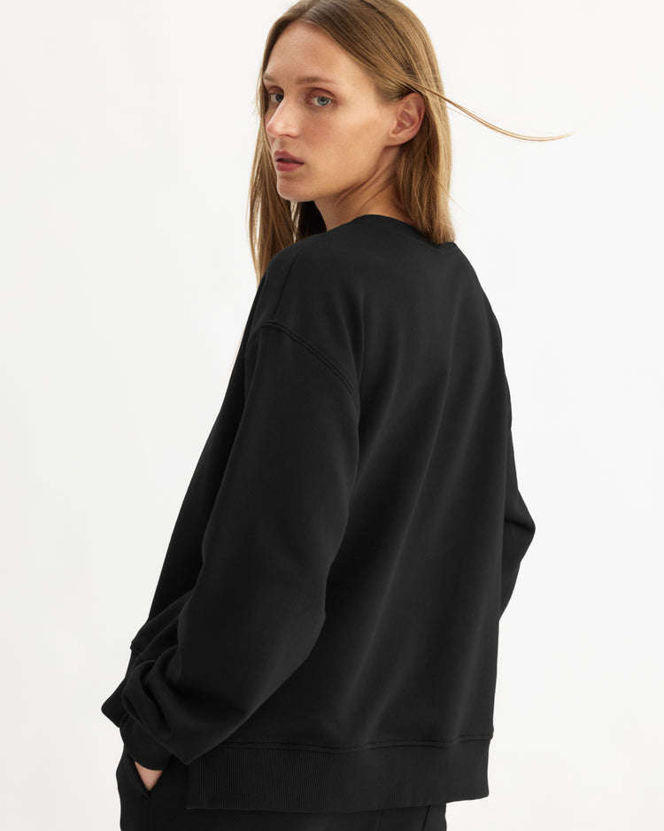 A person with long, light brown hair stands sideways against a plain white background, looking back over their shoulder while wearing the Arlo Mott Delancey Crew Neck Sweatshirt in Black.