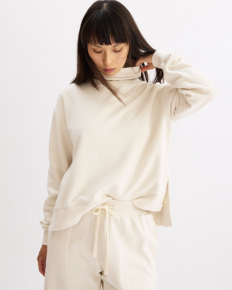 A woman with straight dark hair and bangs wears the Arlo Mott Delancey Crew Neck Sweatshirt in Chalk, paired with matching drawstring pants, looking down and touching her neck against a plain white background.