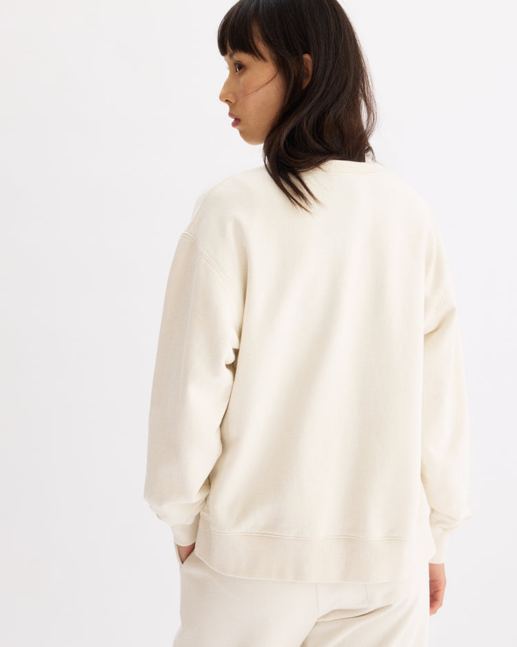 A person with medium-length dark hair, wearing the Arlo Mott Delancey Crew Neck Sweatshirt in Chalk, stands mostly with their back to the camera and looks to the side against a plain white background.