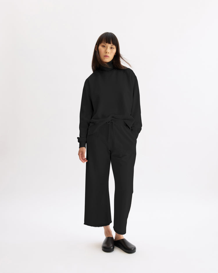A person with straight dark hair wears Arlo Mott’s Kane Cropped Wide Sweatpants in Black, a black long-sleeve top, and black slip-on shoes, standing against a plain white background and looking at the camera.