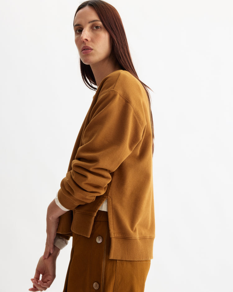A woman with long brown hair stands against a white background, wearing the Arlo Mott Delancey Crew Neck Sweatshirt in Cinnamon and a matching skirt with side buttons. She gazes over her shoulder with a neutral expression.
