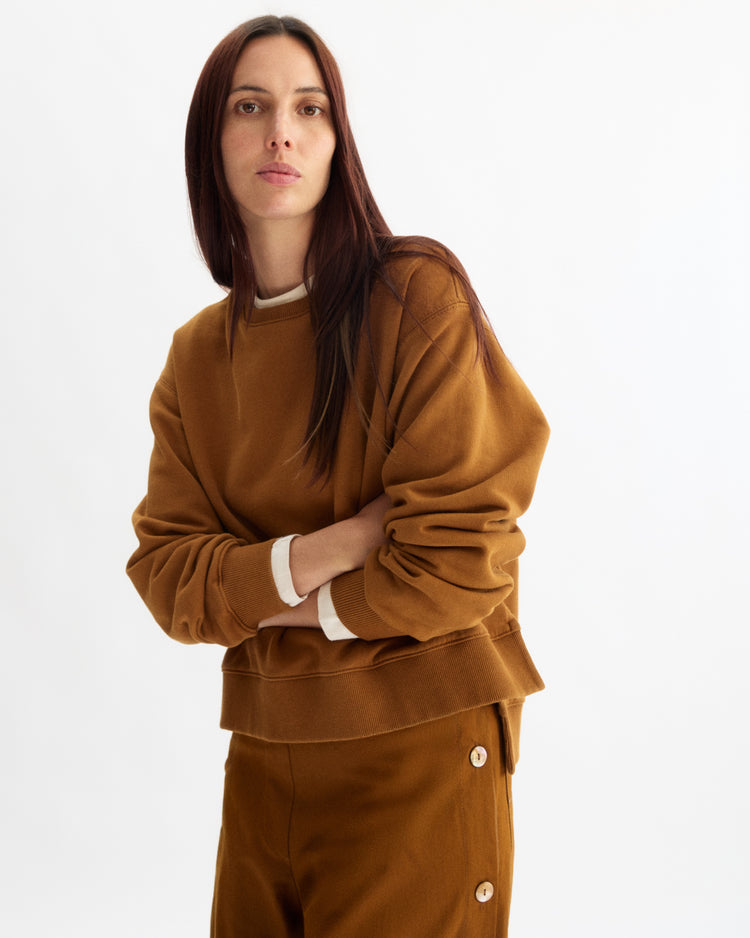 A person with long, straight brown hair models the Arlo Mott Delancey Crew Neck Sweatshirt in Cinnamon, paired with matching pants featuring buttons, posing against a plain white background.