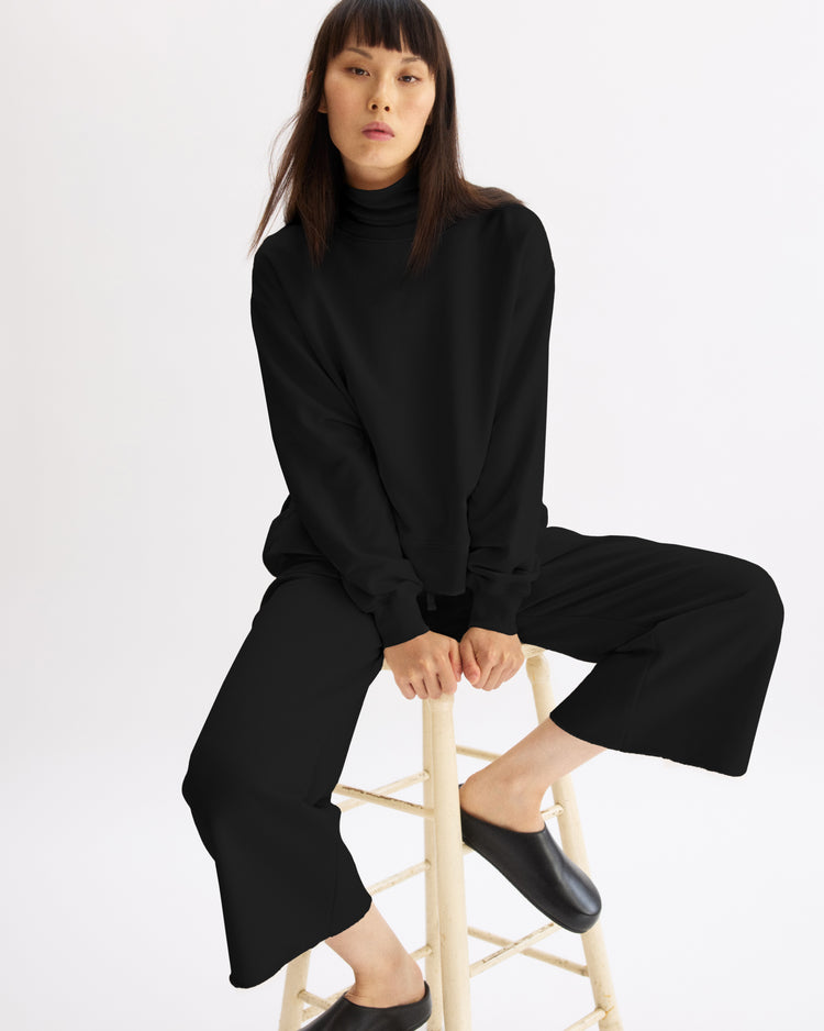 A woman with straight dark hair and bangs sits on a wooden stool against a plain white background, wearing Arlo Mott’s Kane Cropped Wide Sweatpants in Black, a black turtleneck, and black slip-on shoes.