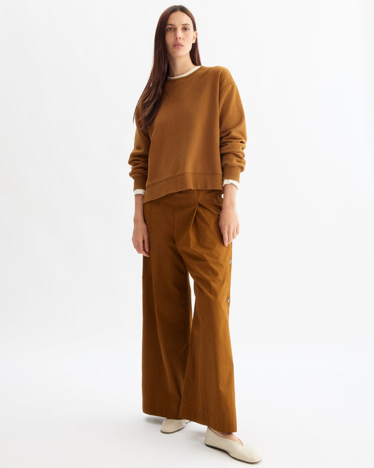 A woman with straight, long brown hair and a neutral expression wears the Arlo Mott Delancey Crew Neck Sweatshirt in Cinnamon against a white background, styled with matching wide-leg pants and white slip-on shoes.
