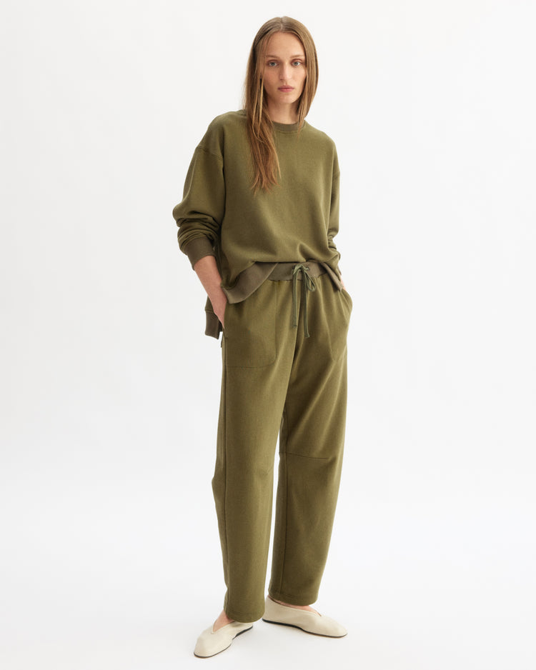 A woman with long, straight hair stands against a white background wearing the Arlo Mott Delancey Crew Neck Sweatshirt in Olive and matching drawstring pants, both in soft Italian terry cloth. She pairs the look with cream-colored slip-on shoes.