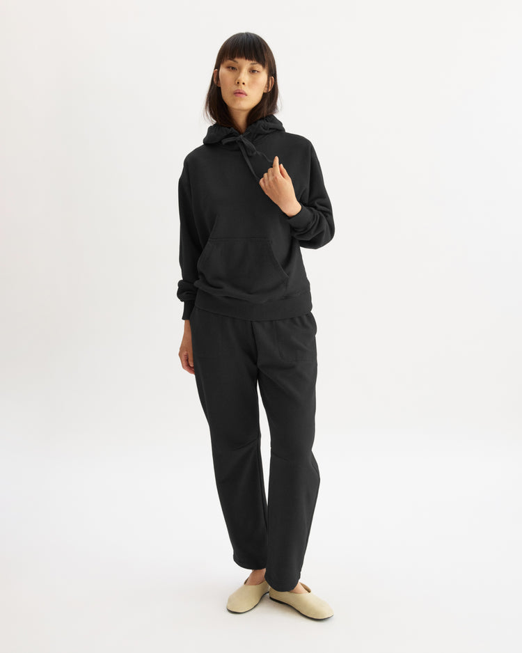 A woman with straight dark hair stands against a white background, wearing Arlo Mott's Baltic Barrel Leg Sweatpants in Black with a black hoodie and light beige slippers. One hand is raised near her collar.