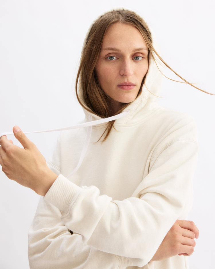 A woman with long, light brown hair wears the Arlo Mott Windsor Hooded Sweatshirt in Chalk, made from cozy Italian terry cloth. She holds one drawstring with her left hand and looks at the camera against a plain white background.