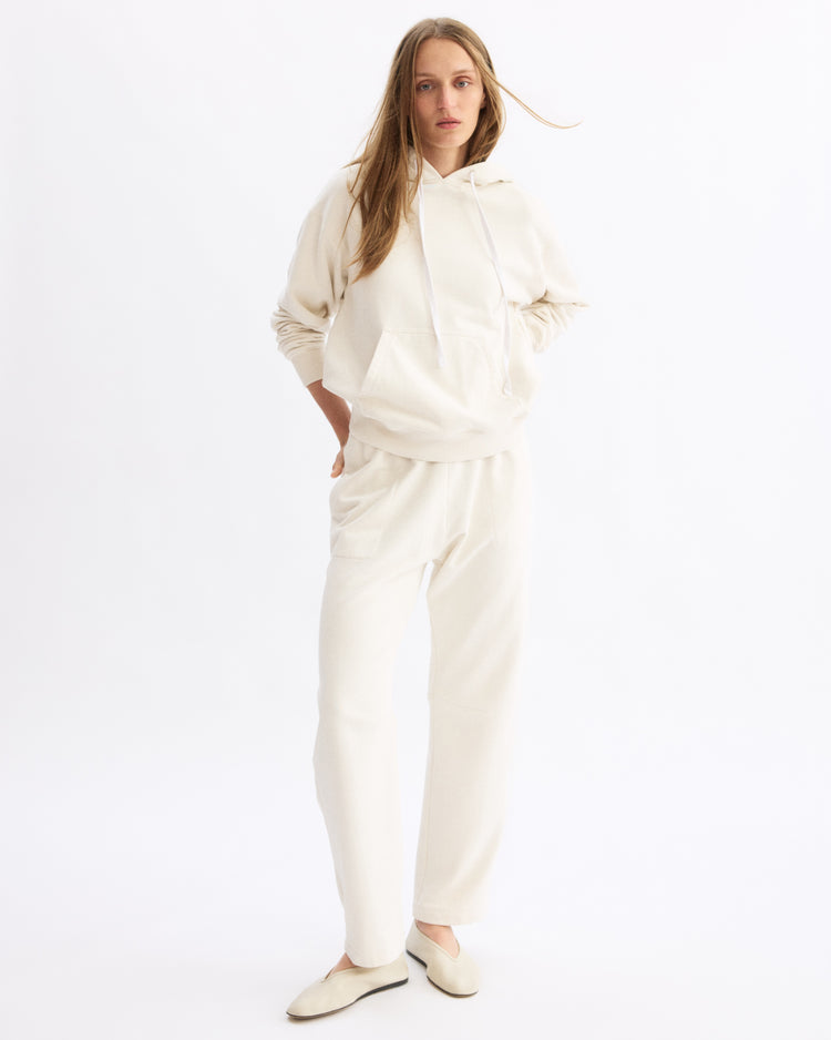 Someone stands against a plain white background wearing the Arlo Mott Windsor Hooded Sweatshirt in Chalk with kangaroo pockets, paired with matching sweatpants and off-white slip-on shoes. Their long, light brown hair is worn down and they look relaxed.