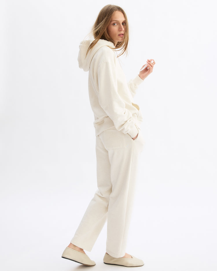 A woman stands against a white background wearing the Arlo Mott Windsor Hooded Sweatshirt in Chalk with matching sweatpants and slip-on shoes. She looks over her shoulder, one hand raised, her long hair loose.