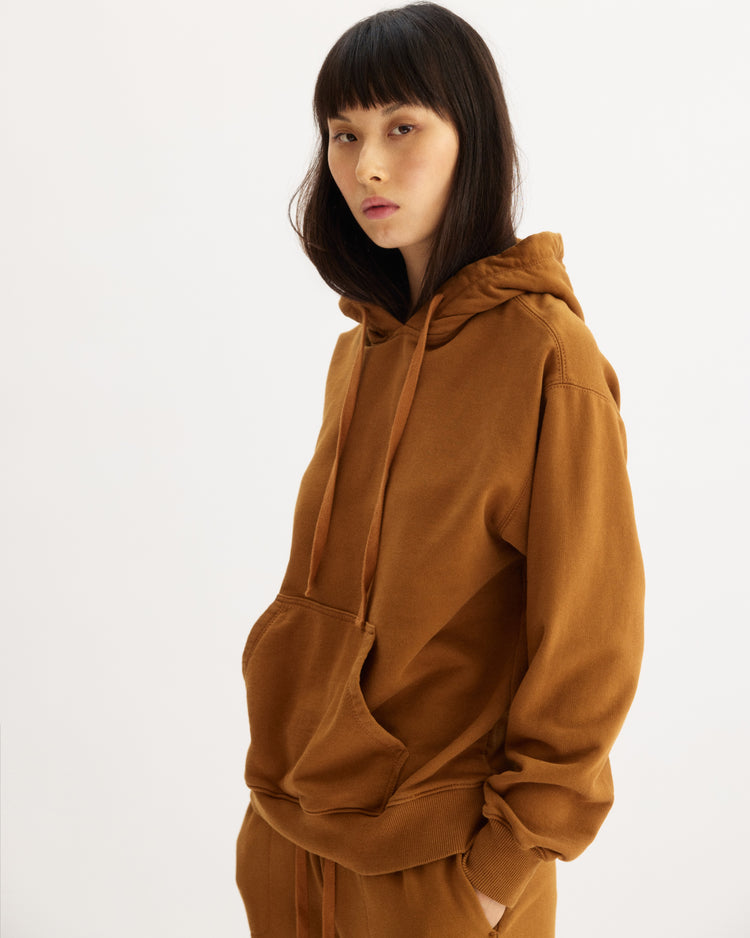 A woman with straight dark hair and bangs wears the Arlo Mott Windsor Hooded Sweatshirt in Cinnamon, featuring kangaroo pockets, standing against a plain white background and looking at the camera with a neutral expression.