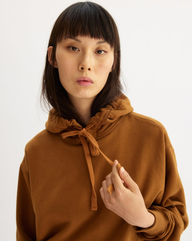 A person with straight dark hair and bangs wears the Arlo Mott Windsor Hooded Sweatshirt in Cinnamon, holding a drawstring in their right hand, standing against a plain light background.