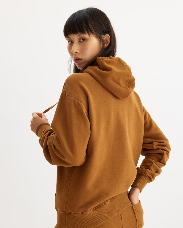 A woman with long dark hair and bangs models the Arlo Mott Windsor Hooded Sweatshirt in Cinnamon, featuring kangaroo pockets, as she looks over her shoulder toward the camera in a neutral, well-lit setting.