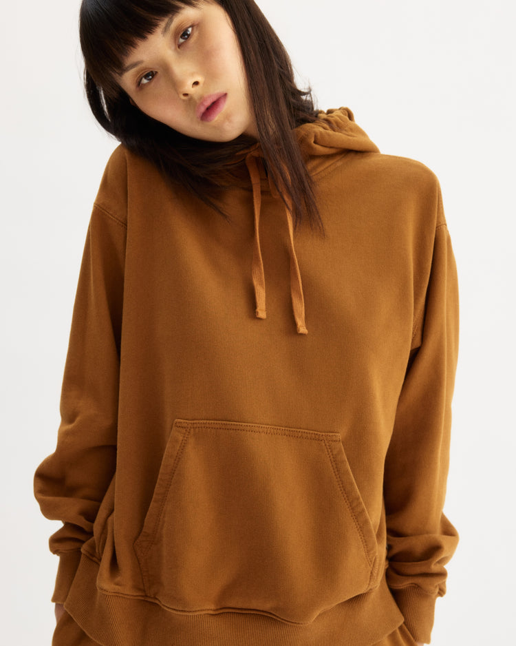 A person with straight dark hair and bangs wears the Arlo Mott Windsor Hooded Sweatshirt in Cinnamon, featuring a loose fit and kangaroo pockets, while standing against a plain white background.