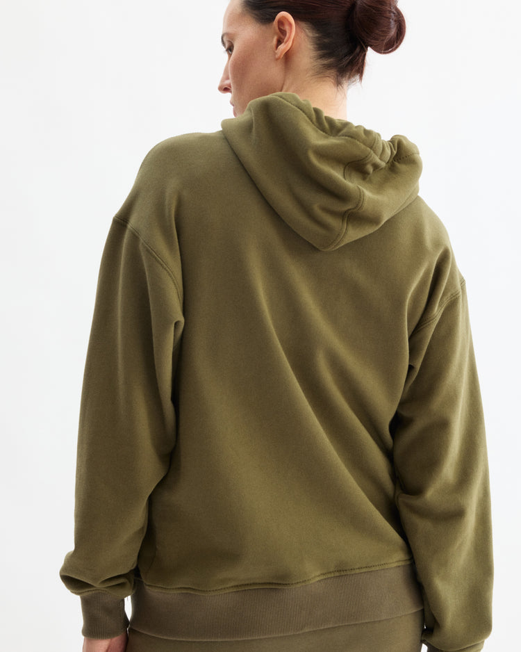 A person with dark hair in a bun wears the Arlo Mott Windsor Hooded Sweatshirt in Olive, featuring plush fabric and kangaroo pockets, facing away from the camera against a plain white background.