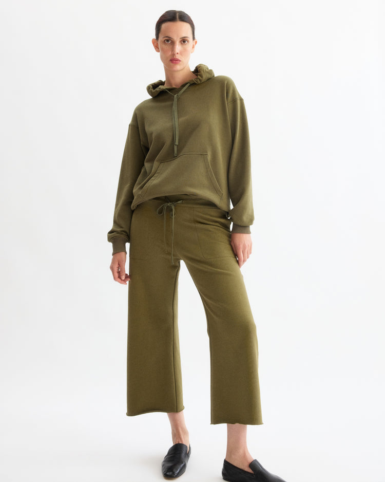 A woman stands against a white background wearing Arlo Mott’s Kane Cropped Wide Sweatpants in Olive with a matching hoodie and black slip-on shoes, facing forward with one hand in her pocket and a neutral expression.