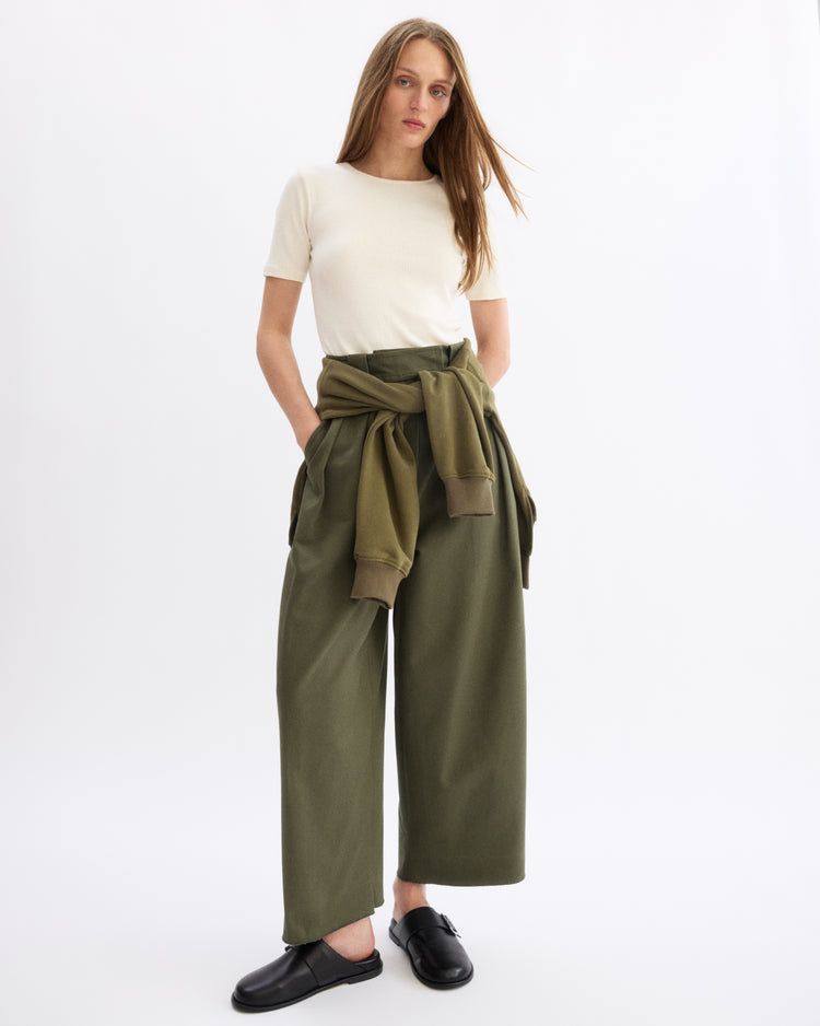 A woman stands against a white background wearing the Arlo Mott Bergen Short Sleeve Tee in Chalk, wide-leg olive green pants, black shoes, and an olive green sweatshirt tied around her waist.
