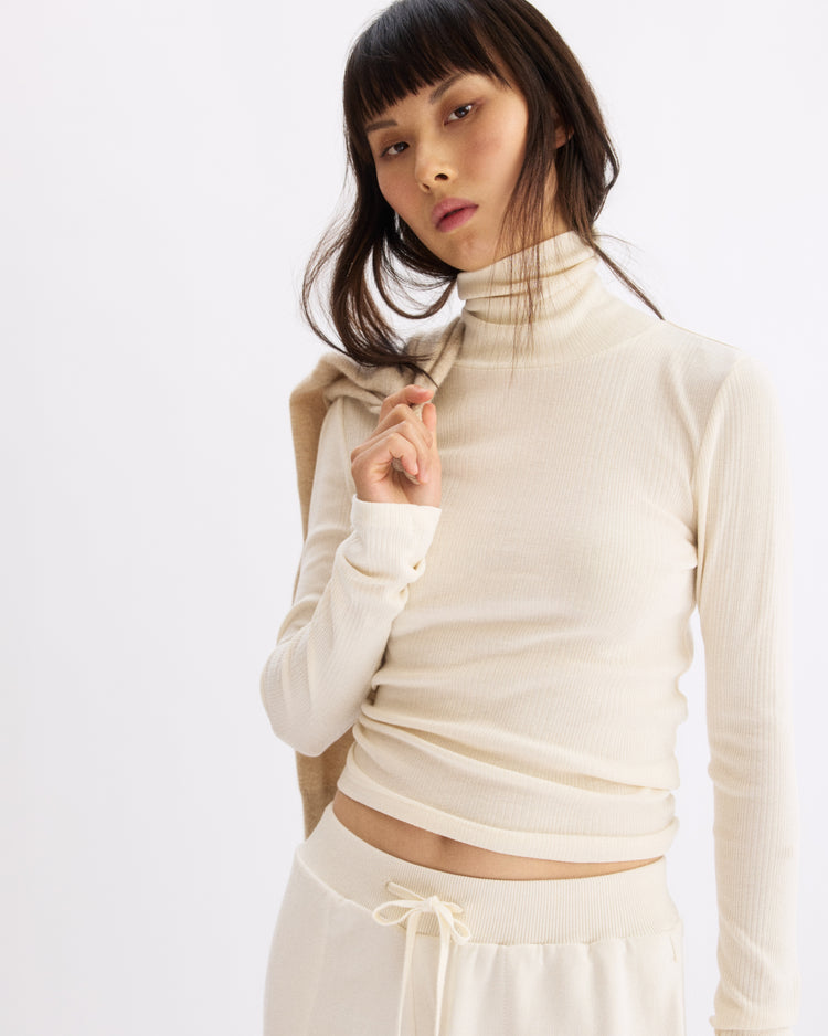 A woman with straight dark hair and bangs wears the Arlo Mott Clinton Long Sleeve Turtleneck in Chalk, holding a light layer over her shoulder against a plain white background.