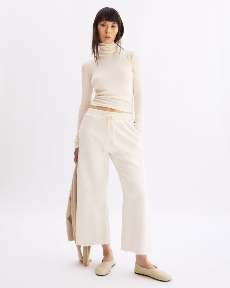 A woman stands against a white background wearing Arlo Mott's Kane Cropped Wide Sweatpants in Chalk with a cream turtleneck and slippers. She holds a beige garment at her side, looking directly at the camera with a neutral expression.