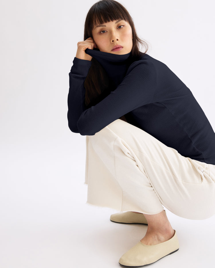 A woman with straight dark hair crouches against a plain background, wearing the Arlo Mott Clinton Long Sleeve Turtleneck in Navy—an ideal layering piece—paired with light cream wide-leg pants and beige slip-on shoes, hand holding the collar to her mouth.