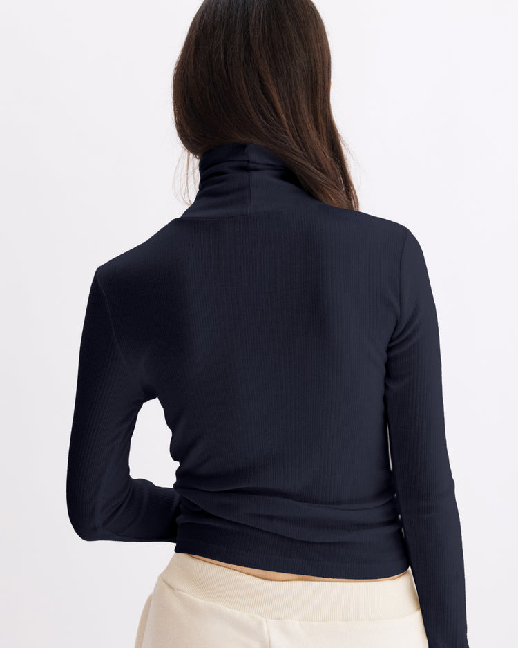 A woman with long brown hair is seen from the back wearing the Arlo Mott Clinton Long Sleeve Turtleneck in Navy and cream-colored pants against a plain light background.