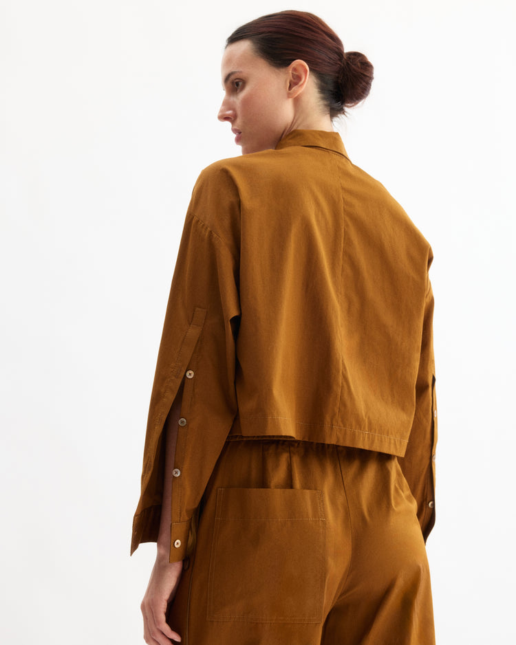 A person with dark hair in a low bun wears the Arlo Mott Hester Boxy Short Button Down in Cinnamon, standing against a plain white background and looking to the side.