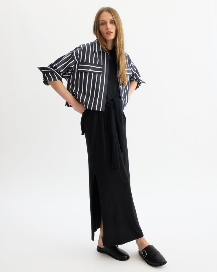 A woman stands against a white background wearing a black ankle-length skirt with a side slit, black top, and the Arlo Mott Hester Boxy Short Button Down in Navy Stripe, paired with black slip-on shoes with a buckle.