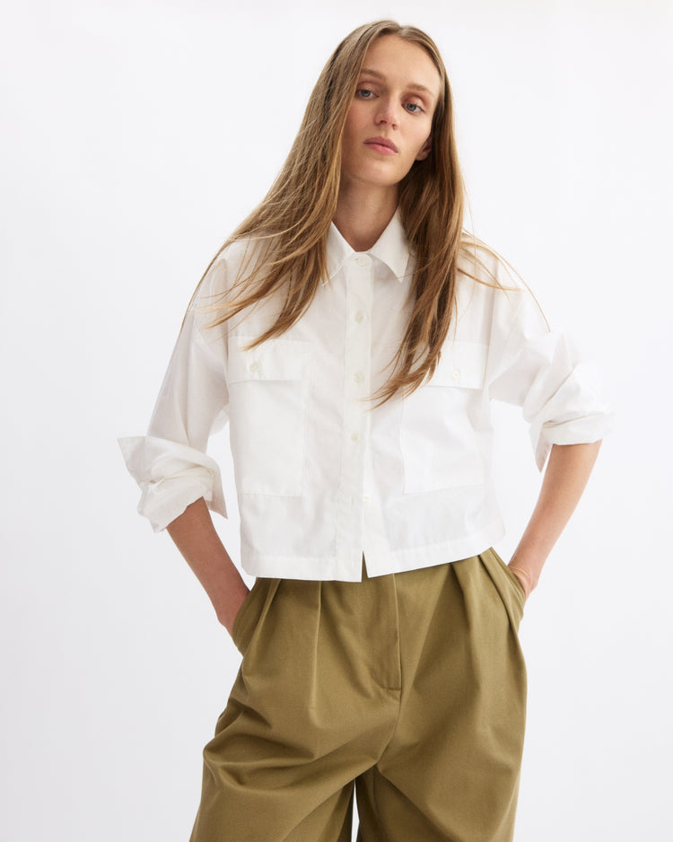 A person with long, light brown hair wears the Arlo Mott Hester Boxy Short Button Down in White and loose khaki pants, standing with hands in pockets against a plain white background.