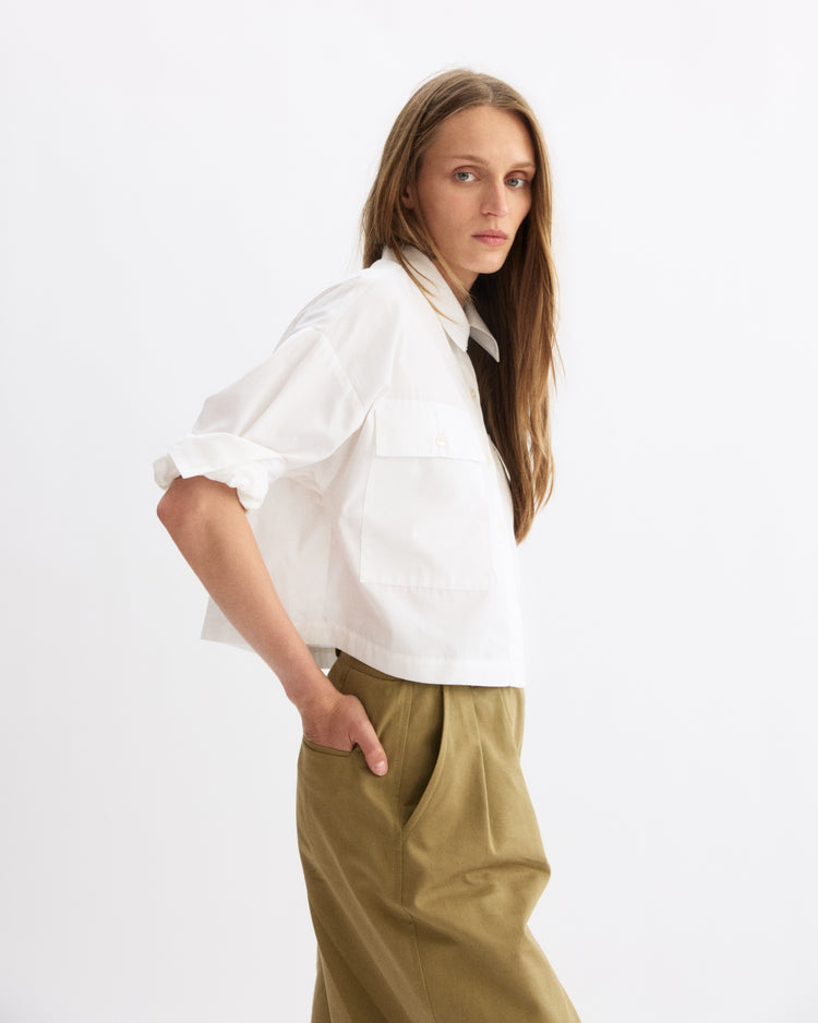 A woman with long, straight hair wears the Arlo Mott Hester Boxy Short Button Down in White and khaki pants. She stands sideways, one hand in her pocket, looking at the camera against a plain white background.