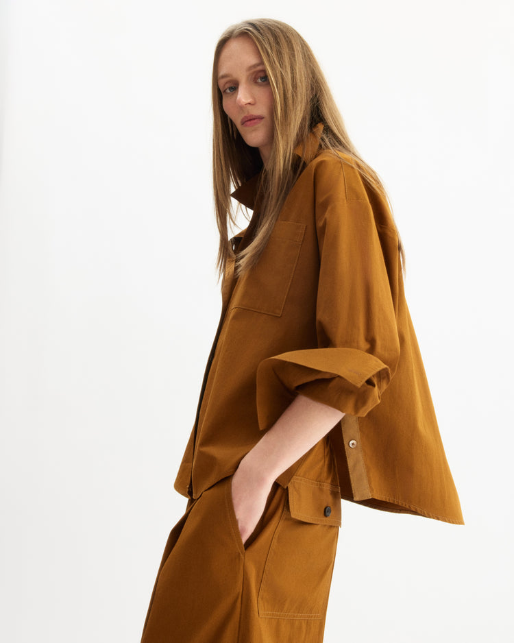 A woman with long straight hair models the Arlo Mott Watts Side Button Shirt in Cinnamon, featuring a loose fit and large pockets, paired with matching pants, standing against a plain white background.