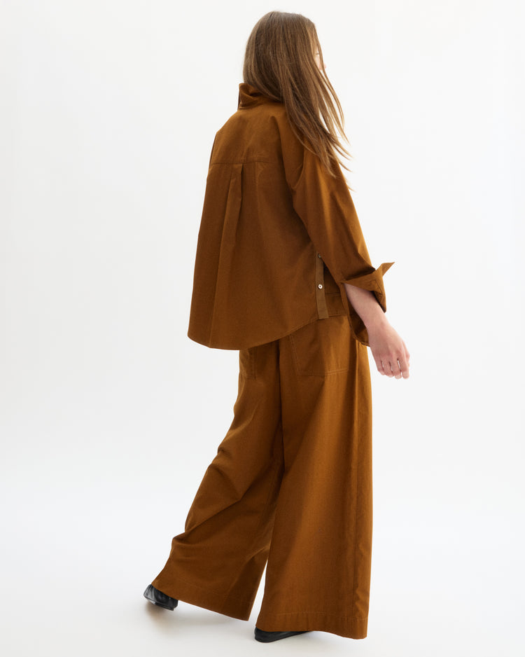 A person with long hair, wearing a loose brown shirt and Arlo Mott's Hudson Pull-on Pants in Cinnamon, made from soft Portuguese cotton with an elastic waistband, stands against a plain white background facing slightly away.