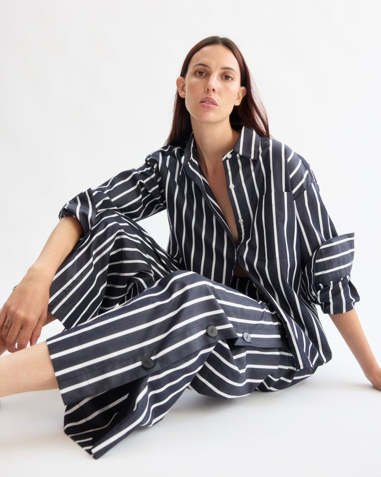 A woman with long dark hair sits on the floor against a white background, wearing Arlo Mott's Baxter Side Button Pants in Navy Stripe, looking directly at the camera with a neutral expression.