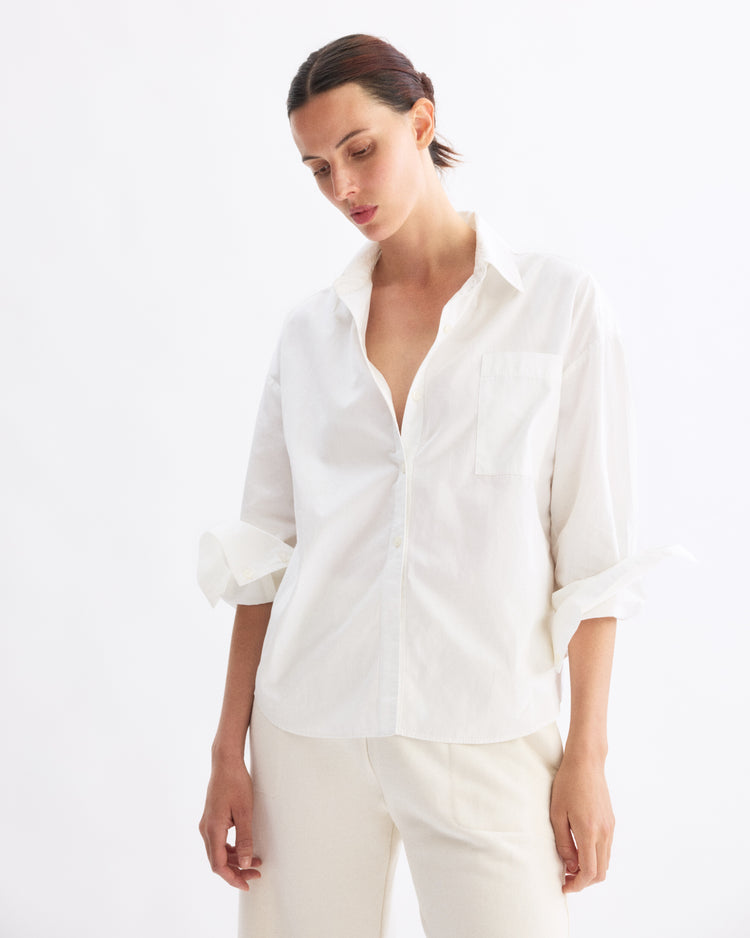 A woman with dark hair pulled back wears the Arlo Mott Watts Side Button Shirt in White with sleeves rolled up and matching white pants, standing against a plain white background.