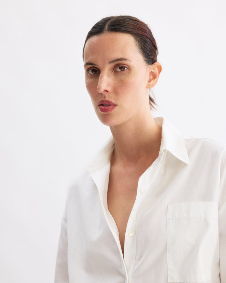 A woman with fair skin and dark hair pulled back wears the Arlo Mott Watts Side Button Shirt in White. She looks slightly to the side with a neutral expression against a plain, light background.