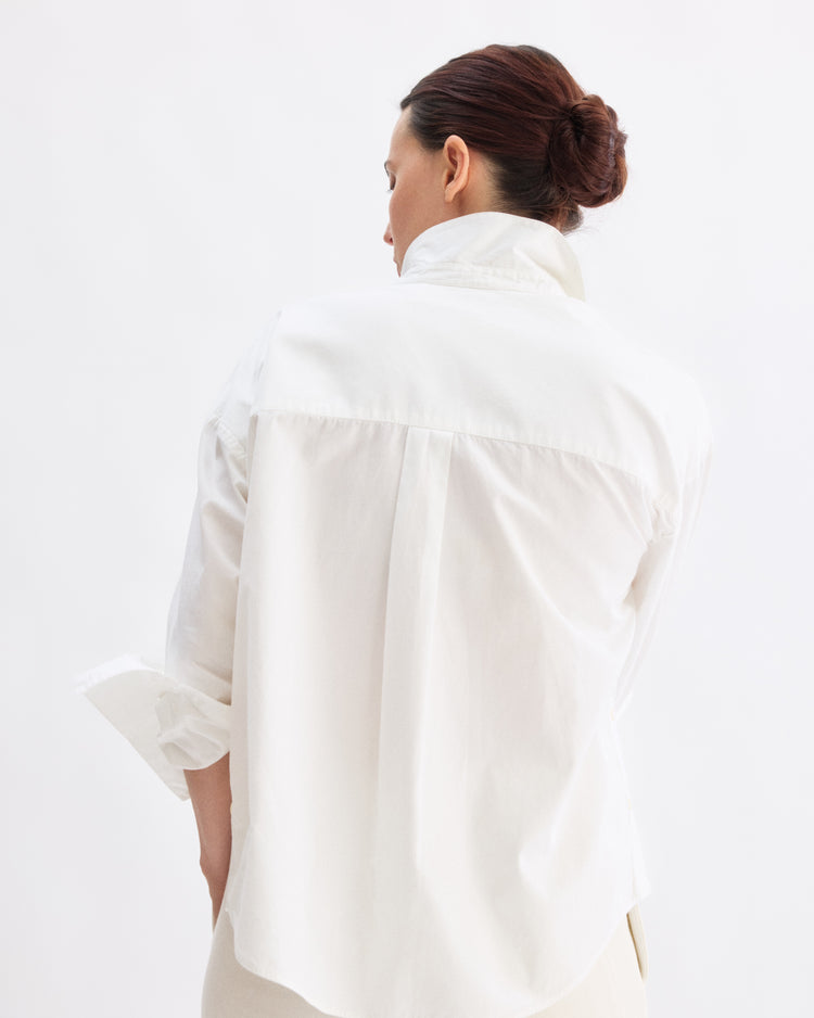 A woman with dark hair in a neat bun, tied with a grosgrain ribbon, faces away from the camera wearing the Arlo Mott Watts Side Button Shirt in White, sleeves rolled up, against a plain light background.