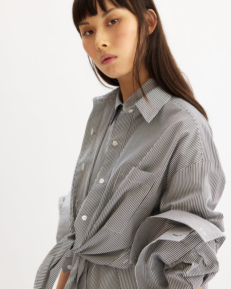 A woman with long dark hair and straight bangs wears the Arlo Mott Watts Side Button Shirt in White Stripe, a loose-fitting button-down made from Portuguese poplin, as she gazes confidently at the camera against a plain background.