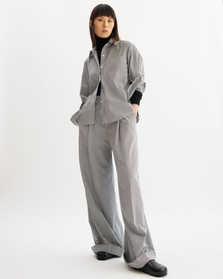 A person stands against a white background, wearing the Arlo Mott Watts Side Button Shirt in White Stripe with matching wide-leg pants. They have straight dark hair with bangs and black shoes.